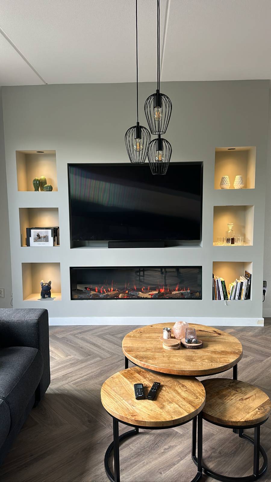 Modern woonkamer met een ingebouwde elektrische haard onder een grote flatscreen-tv, omringd door verlichte nissen met decoratieve items, zoals boeken, glazen en planten. De ruimte bevat ook een set ronde houten salontafels en een donkergrijze bank, met een stijlvol lichtgrijs wanddesign.